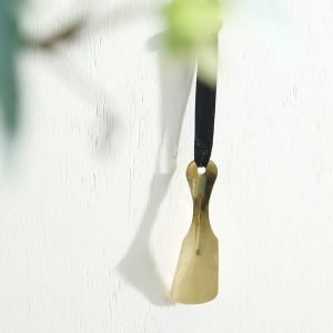 黃銅製鞋拔掛在白色牆上的照片,展示其光亮的金屬質感和精美設計。 Picture of a brass shoehorn hanging on a white wall, showcasing its shiny metal texture and elegant design.