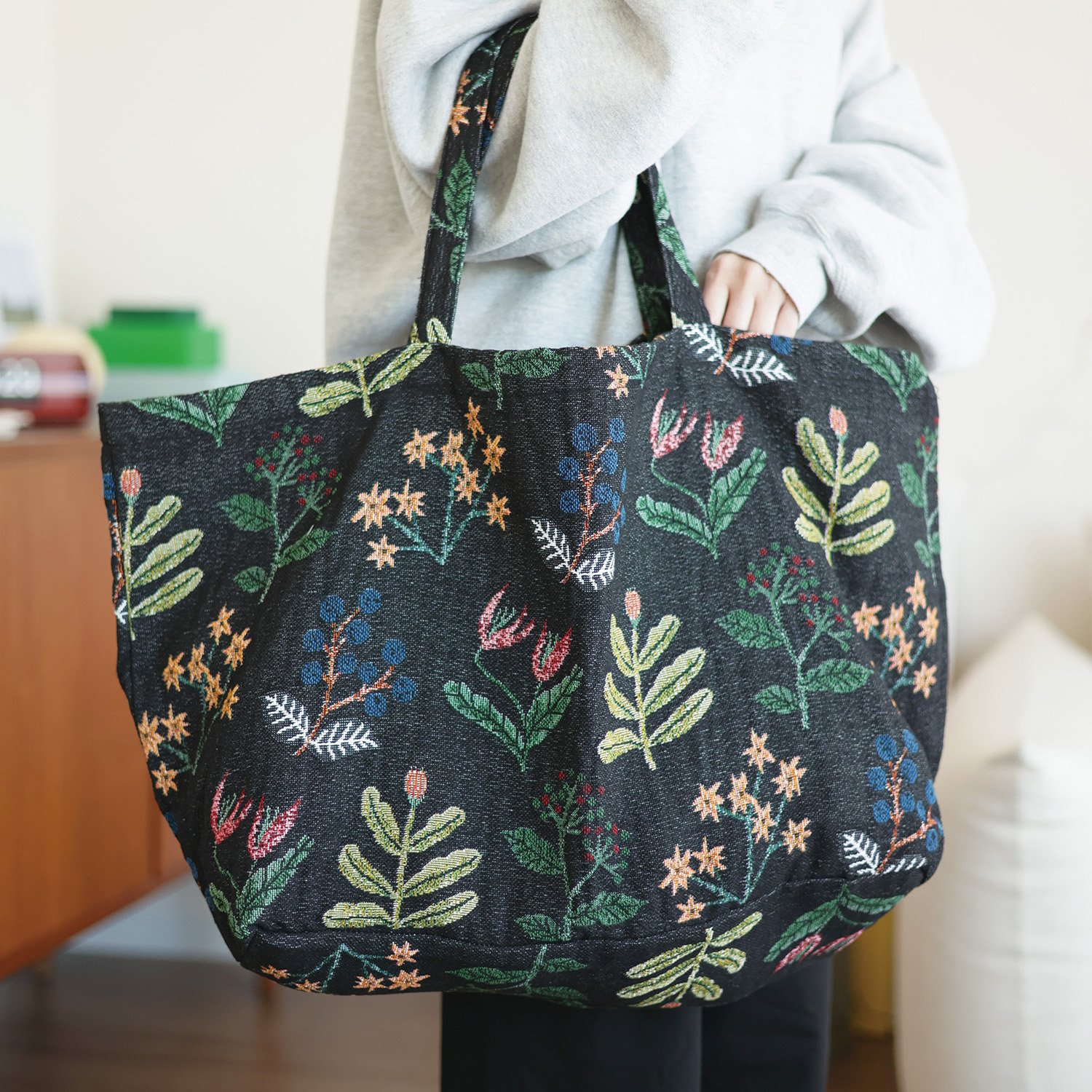 展示一隻手挽著Miyuki 松尾黑色花朵大袋的照片，清晰顯示袋子的正面設計和細節. An image showing one hand holding the &quot;Miyuki 松尾黑色花朵大袋,&quot; clearly displaying the front design and details of the bag.