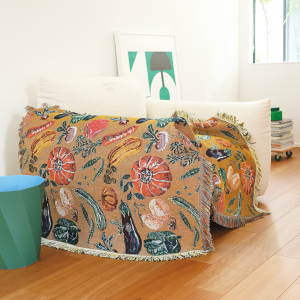 展示Nathalie Lete 蔬菜圖案掛毯地毯的圖片，展開並放置在沙發的扶手上，背景為舒適的室內環境. An image showing the vegetable design of the Nathalie Lete tapestry rug spread out and placed on the armrest of a sofa, set in a cozy indoor environment.