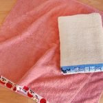 一條貓咪圖案毛巾放在桌子上,位於蘋果設計毛巾之上,左上角有K'S Cottonhouse標誌. A cat pattern towel placed on a table, resting on top of an apple design towel, with the K'S Cottonhouse logo in the upper left corner.v