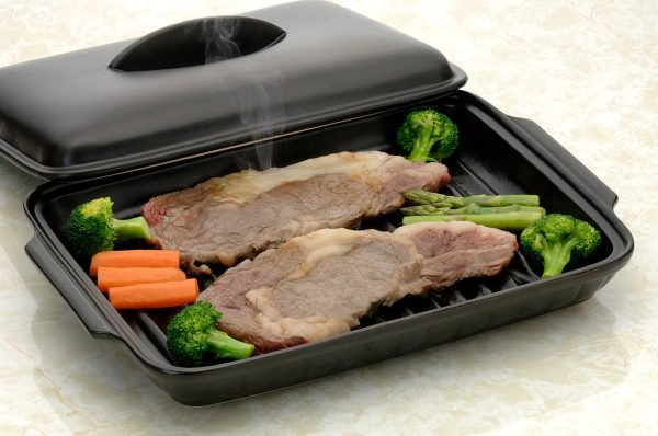 日本製造陶瓷烤盤內含牛排和蔬菜，耐熱蓋擱放在盤邊. A Japanese ceramic baking tray with steaks and veggies inside, with the heat-resistant lid resting on the edge of the tray.