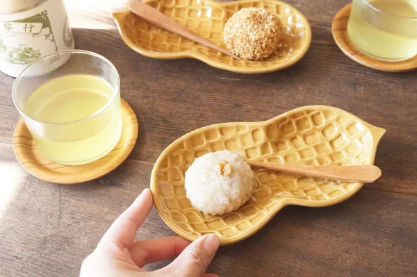 餐桌視角,兩個手指握住盛有食物的花生造型盤,旁邊有另一個食物盤和兩杯飲料 - View of a dining table with two fingers holding a peanut-shaped plate of food, another food plate, and two glasses.