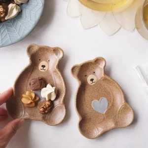 兩個手指握住盛有餅乾的Yoshizawa 益子燒泰迪熊小盤，旁邊有一個空盤 - Two fingers holding a Yoshizawa Mashiko-style teddy bear small plate with cookies, with an empty plate beside it.