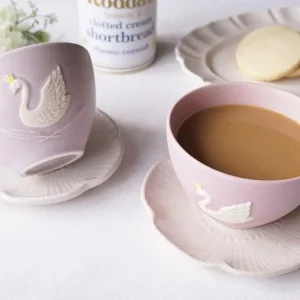 桌上兩個Yoshizawa 益子燒紫色皇冠天鵝杯，一個有飲料，另一個側放在碟子上，旁邊有餅乾。 / Two Yoshizawa Mashiko-style purple crown swan cups on a table, one with a drink and the other lying on a saucer, with cookies nearby.