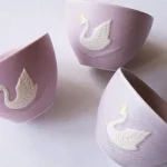 三個Yoshizawa 益子燒紫色皇冠天鵝杯擺放在桌上，展示側面的天鵝圖案，色調各異，展現手工製作的獨特性。 / Three Yoshizawa Mashiko-style purple crown swan cups on a table, showcasing the swan pattern on the sides, each with unique color tones reflecting their handmade nature.