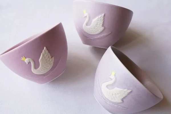 三個Yoshizawa 益子燒紫色皇冠天鵝杯擺放在桌上，展示側面的天鵝圖案，色調各異，展現手工製作的獨特性。 / Three Yoshizawa Mashiko-style purple crown swan cups on a table, showcasing the swan pattern on the sides, each with unique color tones reflecting their handmade nature.