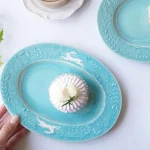 兩個Yoshizawa 益子燒綠色兔子花卉橢圓餐盤放在桌上，前面的盤子上有一個蛋糕，並展示一隻手握住盤子 - Two Yoshizawa Mashiko-style green rabbit floral oval plates on a table, with the front plate showing a cake and a hand holding it.