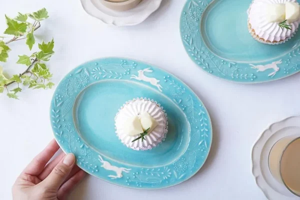 兩個Yoshizawa 益子燒綠色兔子花卉橢圓餐盤放在桌上，前面的盤子上有一個蛋糕，並展示一隻手握住盤子 - Two Yoshizawa Mashiko-style green rabbit floral oval plates on a table, with the front plate showing a cake and a hand holding it.