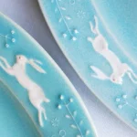 兩個Yoshizawa 益子燒綠色兔子花卉橢圓餐盤的特寫，展示邊緣的兔子和植物圖案 - Close-up of two Yoshizawa Mashiko-style green rabbit floral oval plates, showing the rabbit and plant patterns on the edges