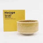 Heuge 抹茶碗 黃瀬戶與包裝盒擺放在一起,展示碗的設計和其精美的包裝。 / Heuge matcha bowl in yellow Seto displayed with its packing box behind, showcasing the bowl's design and elegant packaging.