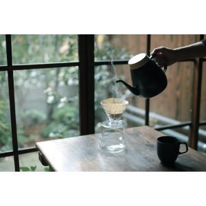 右手拿着黑色的日本製 Ovject 搪瓷咖啡壺，將水倒進木枱上的杯，背景可見窗外的景色