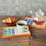 山茶花與手菊款的日本製SATOMIMI 咖啡托盤放在木枱上，山茶花款上放着玻璃咖啡壺，手鞠款托盤上放着一杯咖啡，枱上另有一個碟子裝着牛角麵包放在托盤旁邊