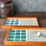 山茶花與手菊款的日本製SATOMIMI 咖啡托盤 放在木枱上的側面圖，前面的手鞠款托盤上放着一隻手錶。