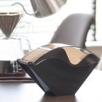 日本製 下村咖啡濾紙收納盒黑色款放在枱面上的正面圖，背後有人拿着咖啡壺正在沖咖啡的朦朧影像