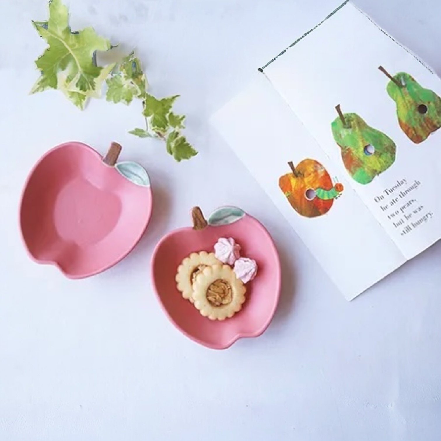 兩個紅色Yoshizawa 益子燒蘋果小盤放在枱上，旁邊放咗一本圖畫書，書上有蘋果及啤梨的圖案