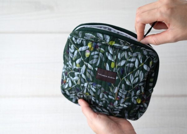 雙手拿着miyaco nishio 有機棉絎縫化妝包 –橄欖款，可以看到袋子的正面，正在拉開拉鍊的白色木板背景圖