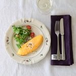 Studio M' Rosier 圓餐盤 24cm 款裝着食物放在木枱上的俯視圖,餐盤旁邊放着紫色餐巾與刀叉。