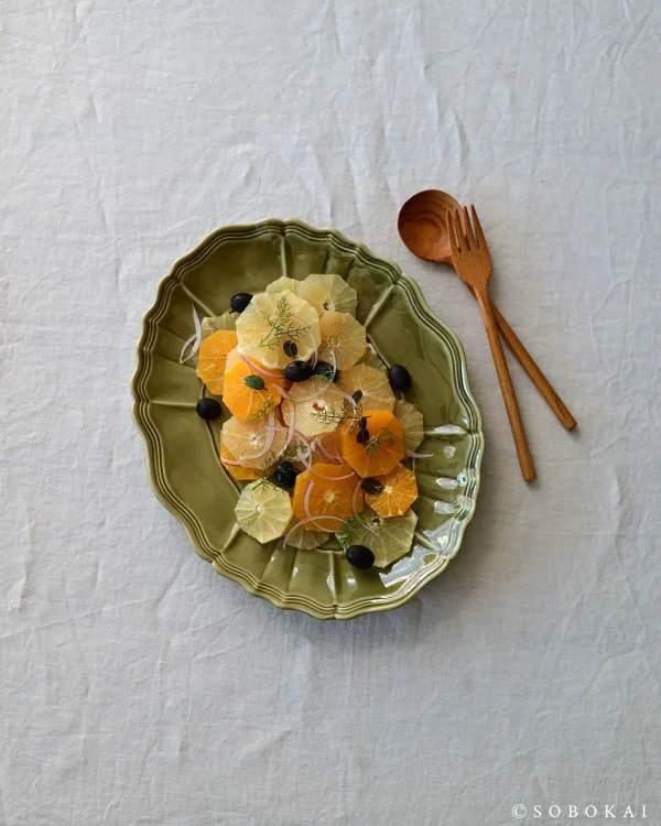 橄欖綠色款 STUDIO M' Sobokai Communus 饗宴浮雕盤 (32.5cm) 裝着食物放在木餐桌上的俯視圖，旁邊有木匙羹與叉子。