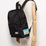 Miffy 黑色背包的前袋上印有色彩繽紛的卡通兔子圖案，展示在素色背景下的木質手臂模特兒身上。.